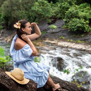 Beautiful girl in a blue dress looks at a Maui waterfall sitting on a rock