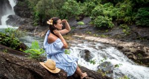 Beautiful girl in a blue dress looks at a Maui waterfall sitting on a rock